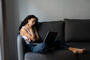 Thoughtful freelance woman sitting on sofa with laptop, touching her neck, feeling tired or stressed during remote work at home in natural light from window.