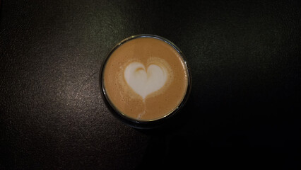 Top-down view of a creamy latte with heart-shaped latte art in a glass cup, set against a dark textured surface. A minimalistic and inviting composition perfect for coffee and café themes.