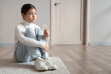 Sad girl with house figure sitting on carpet in hallway. Adoption concept