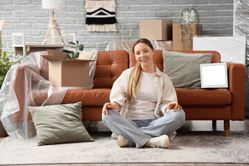 Obraz premium Young woman sitting near sofa with blank laptop in room on moving day