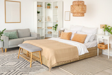 Interior of light bedroom with cozy bed, sofa and shelving unit
