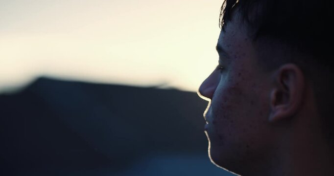 A young man with acne on his face is looking down backlit by the sunset setting sun. Concept of sadness and loneliness, cinematic isolated shot