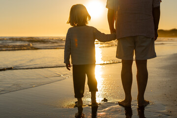Back view of barefoot grandfather and grandson holding hands, watching the sunset over the ocean on the beach, symbolizing love, family bond, and peaceful generational connection.