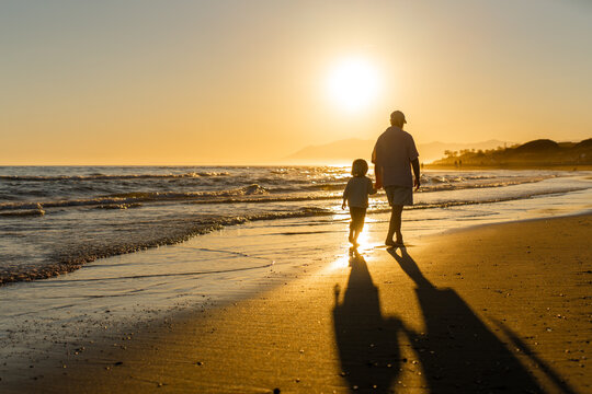 Grandfather and grandson walking barefoot along the beach at sunset, holding hands and casting long shadows in golden evening light by the sea.