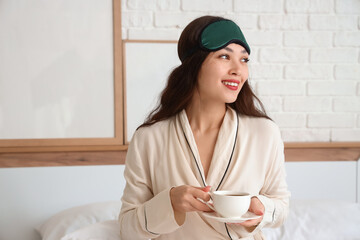 Beautiful young woman with sleep mask and cup of coffee sitting on bed at home