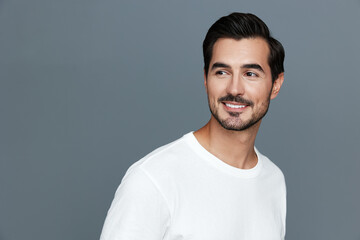 Smiling man in white t-shirt looking to the side with a charming smile, standing against a grey background. Portrait of happiness and confidence.