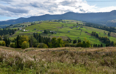 Obraz premium Idyllic rural scene featuring green grassy hills, scattered trees, and small country houses against a background of mountains under a cloudy sky. Carpathian Mountains, Ukraine