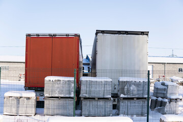 Two truck trailers in snowy warehouse yard with pallets and fence on clear winter day
