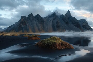Misty rugged mountain peaks with sharp ridges overlooking black sand beach and moss-covered rocks under cloudy sky