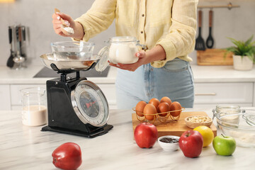 Woman adding flour into bowl on mechanical kitchen scale at white marble table indoors, closeup