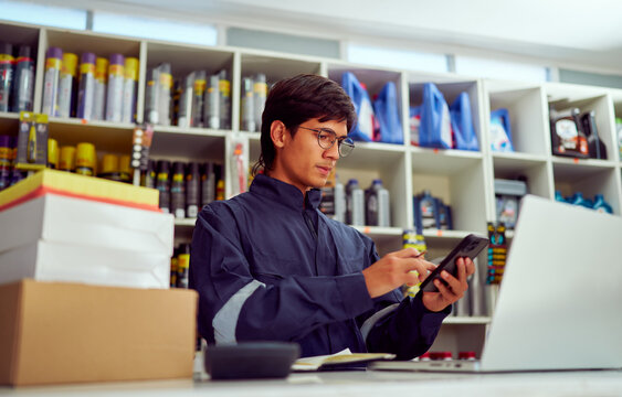 Shop assistant calculating inventory using smartphone and laptop in auto parts store