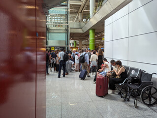 Crowded waiting area at Spanish modern airport terminal. Gathered seated passengers, standing travelers and people with luggage. Busy travel hub, delays and cancelations stress experience. 
