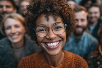 A joyful young woman with curly hair and glasses is smiling broadly in a diverse crowd. The gathering appears lively and festive, suggesting an outdoor celebration or community event.