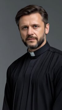 Portrait of mature man in black cleric shirt with a white collar on gray backdrop looking at camera with gray beard and graying hair