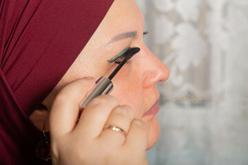 Muslim woman in red hijab sits in room at mirror and applies make-up mascara close-up