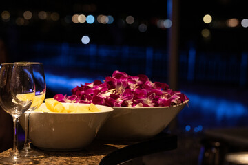 Elegant nighttime setting with vibrant purple orchid leis in a bowl, fresh pineapple slices, and wine glasses, set against a soft city light bokeh—perfect for tropical celebrations and events.