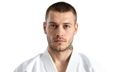 A serious-looking young man with short brown hair and blue eyes, wearing a white martial arts uniform, is shown from the chest up against a white background