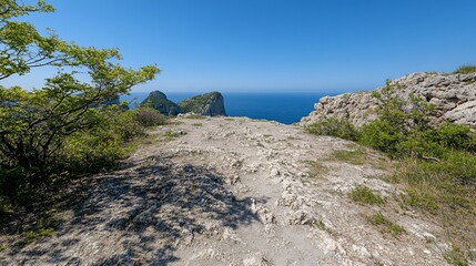 Obraz premium A sun-drenched coastal path winds along a rocky clifftop, overlooking a tranquil azure sea and distant islands under a clear blue sky