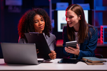 Two confident businesswomen plan strategies while using laptops in a night meeting where files are shared with reports late at night at modern office working on startup company projects.