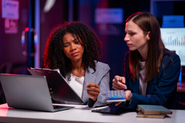Two confident businesswomen plan strategies while using laptops in a night meeting where files are shared with reports late at night at modern office working on startup company projects.