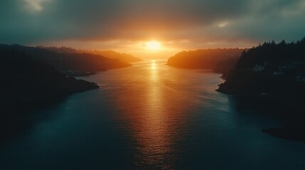 Fototapeta premium Coastal sunset, tranquil waterway. Misty, dramatic skies meet a serene body of water, highlighting a golden sunset. Silhouetted, forested hills flank the calm expanse