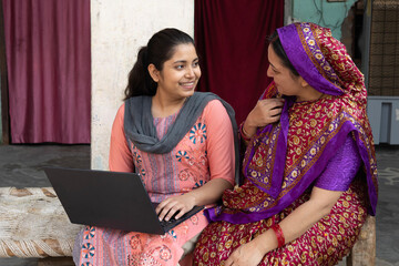 Indian Rural teenager daughter using laptop with mother. Happy villager woman sitting with her...