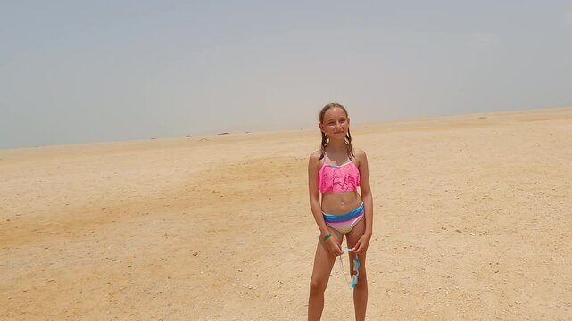 Blonde girl in pink bikini standing on expansive, deserted sandy beach, holding blue swimming goggles while gazing toward horizon under sunny sky