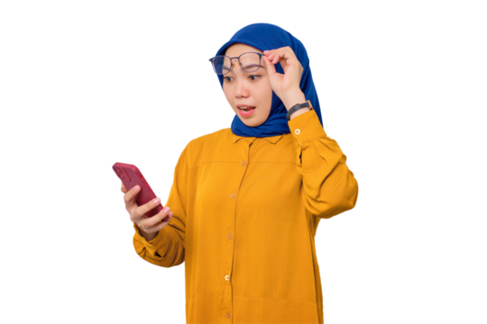 Shocked young Asian Muslim woman dressed in orange shirt looking at mobile phone screen, taking off glasses with opening mouth, reacting to great sale offers isolated on transparent background
