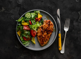 Balanced diet lunch - baked chicken breast and fresh vegetable salad on a dark background, top view