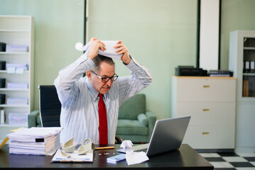 Senior businessman rips paperwork in rage at office desk. Workplace stress, anger, chaos, and...