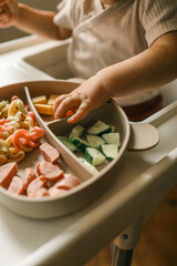 A toddler’s chubby hands grasp colorful pasta and cheese cubes from a divided plate during mealtime