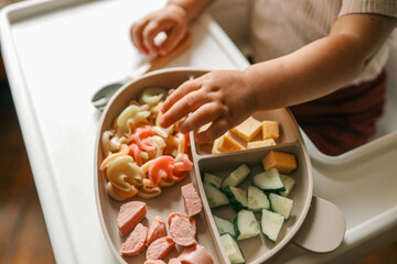 A toddler’s chubby hands grasp colorful pasta and cheese cubes from a divided plate during mealtime