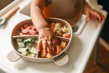 A toddler’s chubby hands grasp colorful pasta and cheese cubes from a divided plate during mealtime