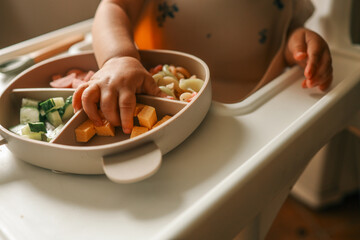 A toddler’s chubby hands grasp colorful pasta and cheese cubes from a divided plate during...