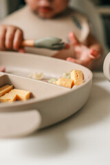 A toddler’s chubby hands grasp colorful pasta and cheese cubes from a divided plate during mealtime
