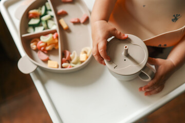 Chubby baby hands gripping a toddler straw cup lid above a divided plate filled with pasta and cheese in a highchair during mealtime
