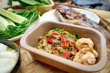 A traditional Thai meal is beautifully arranged on a round bamboo tray. At the center is a perfectly grilled whole fish, served alongside a variety of fresh vegetables, cucumbers, and boiled eggs.