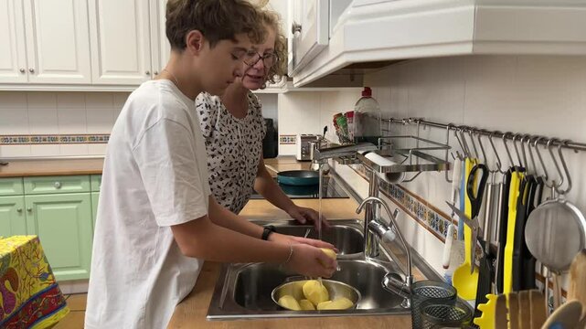 Teenage boy washing potatoes at kitchen sink while his mother explains something to him affectionately