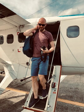 Man stepping off small plane with camera and backpack. Casual travel moment on airport tarmac during vacation.