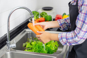 Close-up washing fresh carrots under running water in a kitchen sink, with green lettuce and colorful vegetables in the background, promoting healthy eating, food hygiene, clean cooking preparation