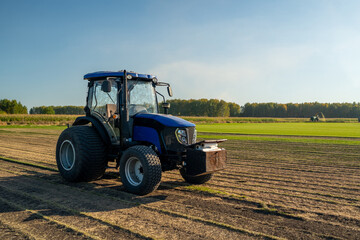 Fototapeta premium A Blue Tractor is Actively Working on a Lush Green Field at the Beautiful Sunrise Hour