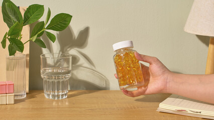 A drug bottle containing capsules full of nutrients is held in the air by someone's hand. On the table was a glass of water. A branch was placed in the bottle. White background with leaves shadows.
