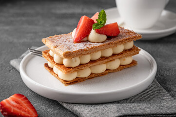 French dessert millefeuille with vanilla cream and fresh strawberries on a white plate on a dark concrete background.