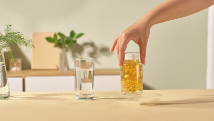 The cylindrical bottle has a transparent shell, white cap and can see the fish oil capsules inside. The yellow pills are filled with vitamins, omegas and other healthy nutrients. Indoor scene.