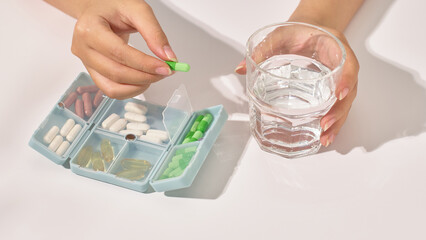 One hand is taking a dietary supplement pill from the personal medicine box, the other hand is holding a glass of water to prepare to take the medicine. The pills have different colors and effects.