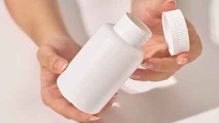 Close-up view of a hand holding unlabeled white open pill bottle, the cap resting in the other hand. The pill box is medium sized, used to hold compressed pills. White background, health supplement.