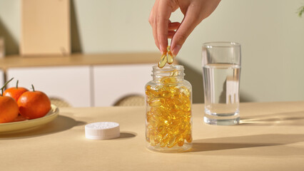 Slender fingers are taking fish oil capsules out of the bottle. The medicine bottle is made of glass, large enough to hold hundreds of pills. The medicine theme photograph, scene in the cozy home.
