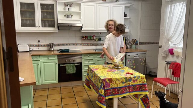 Teenage boy slicing potatoes with mandoline in kitchen while mother gives instructions and does other chores