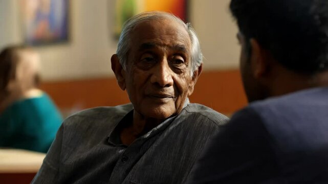 An older South Asian man dressed in a traditional gray kurta engages in a meaningful dialogue with a companion in a cozy cafe filled with warmth