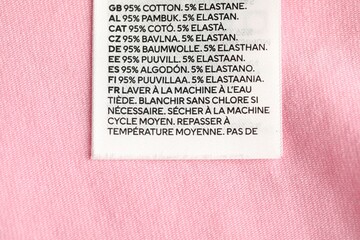 Clothing label with care advice and size on pink garment as background, top view
